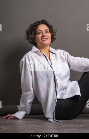 Une jolie femme avec des dents saines parfaitement blanches est assise sur le sol et regarde la caméra. Isolé sur fond gris. Banque D'Images