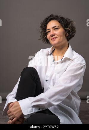 Une jolie femme avec des dents saines parfaitement blanches est assise sur le sol et regarde la caméra. Isolé sur fond gris. Banque D'Images