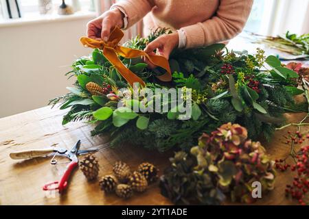 Gros plan de la femme faisant et décorant couronne de Noël festive sur la table à la maison attache Bow fait de ruban Banque D'Images