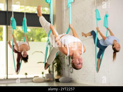 En forme et belle jeune femme suspendue à l'envers tout en pratiquant le yoga aérien pendant le cours de groupe dans le club de fitness Banque D'Images