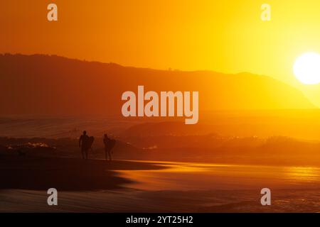 1er décembre 2024. Ehukai Beach Park, Pupukea, Oahu, Hawaï. Deux surfeurs marchent sur la plage au coucher du soleil. Banque D'Images