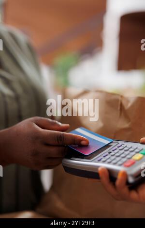 Une femme afro-américaine est vue de près alors qu'elle effectue sans effort un paiement sans contact à la caisse du marché, tenant en toute confiance une carte de crédit et utilisant une machine POS. Banque D'Images