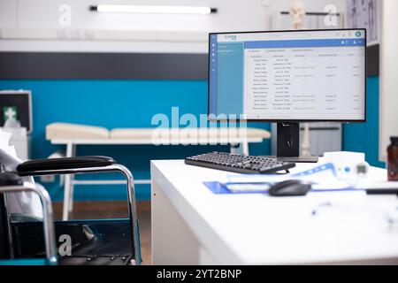 Pupitre vide pour médecin utilisateur de fauteuil roulant avec écran d'ordinateur affichant les dossiers médicaux des patients pour les rendez-vous à venir. Cadre clinique avec outils d'examen et documents médicaux organisés. Banque D'Images