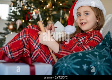 Jolie petite fille heureuse écrivant lettre au Père Noël près de l'arbre de Noël à la maison. Vacances d'hiver. Confortable. Banque D'Images