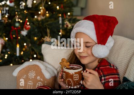 Gros plan jolie petite fille heureuse tenant une tasse de cacao chaud près de l'arbre de Noël à la maison. Vacances d'hiver. Confortable. Banque D'Images