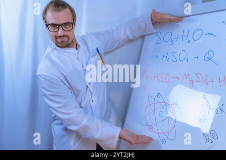 Un scientifique portant des lunettes de sécurité analyse un liquide vert dans un flacon Erlenmeyer, entouré de tubes à essai et d'équipements de laboratoire sur une surface en bois. Che Banque D'Images