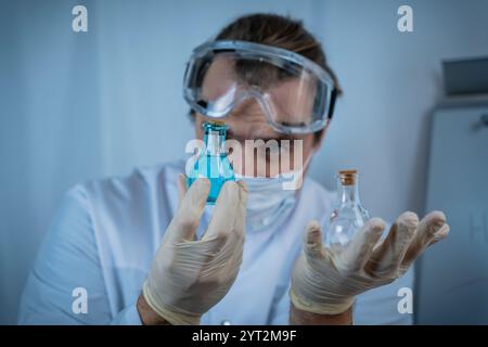 Un scientifique portant des lunettes de protection, des gants et un masque facial analysant soigneusement les flacons contenant des liquides bleus et clairs, en mettant l'accent sur la précision, re Banque D'Images