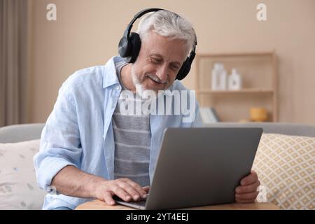 Homme senior avec écouteurs à l'aide d'un ordinateur portable à la table en bois à l'intérieur Banque D'Images