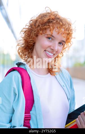 Étudiant joyeux aux cheveux rouges tenant un dossier rouge dans un cadre extérieur lumineux Banque D'Images