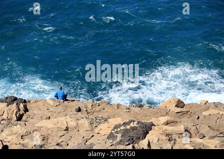 Une scène sereine d'un pêcheur solitaire debout sur une côte accidentée et rocheuse, jetant sa ligne dans le vaste océan. Les vagues s'écrasent contre les rochers. Banque D'Images