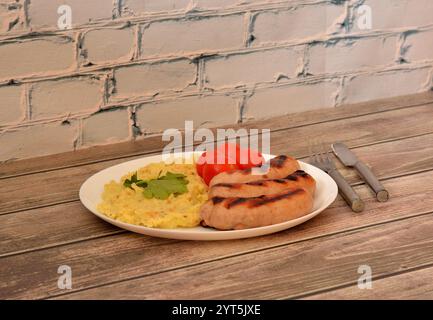 Saucisses grillées avec purée de pommes de terre et tomates, une assiette de délicieux déjeuner sur une table en bois, couverts à proximité. Gros plan. Banque D'Images