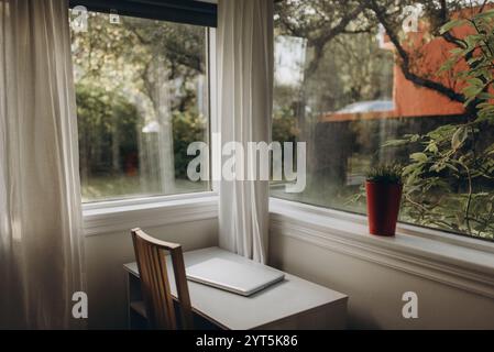 Un coin confortable d'une pièce avec de grandes fenêtres qui laissent entrer la lumière naturelle, un bureau minimaliste et un ordinateur portable ouvert Banque D'Images