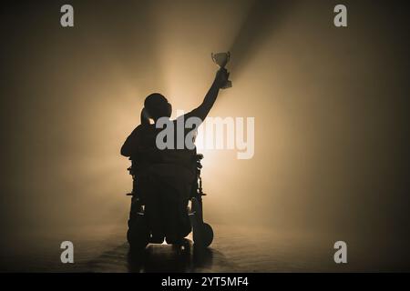 Femme handicapée recevant une coupe de victoire alors qu'elle était assise dans un fauteuil roulant dans une pièce sombre avec un rétroéclairage blanc. Banque D'Images