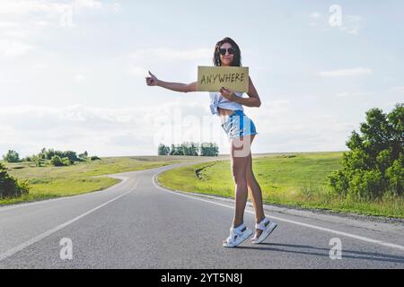 Smiling Girl voyage en auto-stop avec un signe en carton dans ses mains. Espace pour le texte. une fille en lunettes shorts et talons route sur le bord de la route tenant un emp Banque D'Images