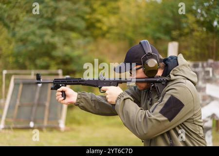 Un homme dans une veste verte tient un pistolet et porte des protections auditives. Il est dans une zone herbeuse avec quelques cibles en arrière-plan Banque D'Images