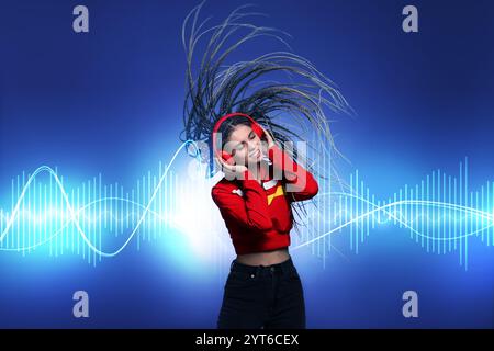 Belle femme avec des écouteurs écoutant de la musique et dansant sur fond bleu. Des ondes sonores derrière elle Banque D'Images