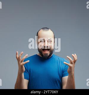 L'homme adulte mi-excité avec la barbe portant un t-shirt bleu crie de joie et de surprise, exprimant des émotions positives sur un fond gris Banque D'Images