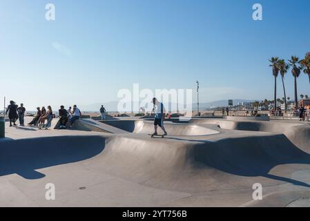 Un skateur se produit sur le béton courbé du Venice Beach Skatepark, avec des spectateurs à proximité. Les palmiers et les montagnes lointaines encadrent la scène. Banque D'Images