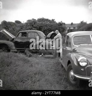 Années 1950, historique, dehors dans un champ herbeux, deux mécaniciens moteurs en salopette travaillant sur une voiture Vauxhall de fabrication britannique de l'époque, roues toutes enlevées, capot relevé. L'un se trouve sous la voiture, l'autre à côté travaillant sur les freins sur le moyeu de roue, Angleterre, Royaume-Uni. Banque D'Images