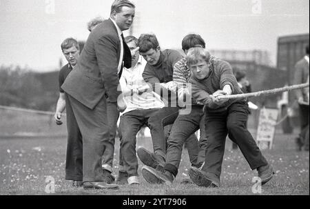 1968, historique, historique, dehors dans un champ à un comté fayre ou fete, une équipe de jeunes gars tirant fort sur une corde dans une compétition de traction de guerre, un manager d'équipe ou un entraîneur dans un costume et cravate debout à côté d'eux donnant des instructions et soutien, Angleterre, Royaume-Uni. Une traction de guerre est une compétition sportive, un test de force qui demande beaucoup d'efforts. Banque D'Images