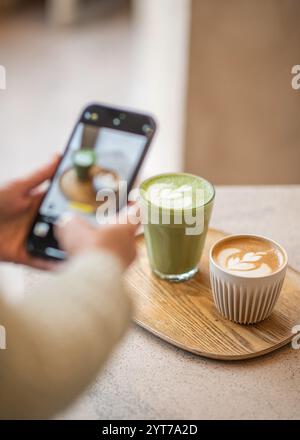 Une main tient un téléphone pour prendre une photo d'un latte et d'une boisson matcha sur un plateau en bois. La scène met en valeur les vibes de café douillettes et les détails du Latte art. Banque D'Images