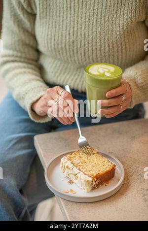 Une scène de café confortable avec une personne dans un pull vert tenant un latte matcha. Sur la table, une tranche de gâteau nappé d'amandes est servie. Banque D'Images