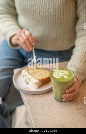 Une scène de café confortable avec une personne dans un pull vert tenant un latte matcha. Sur la table, une tranche de gâteau nappé d'amandes est servie. Banque D'Images