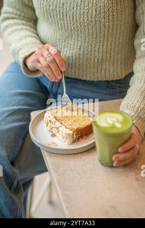 Une scène de café confortable avec une personne dans un pull vert tenant un latte matcha. Sur la table, une tranche de gâteau nappé d'amandes est servie. Banque D'Images