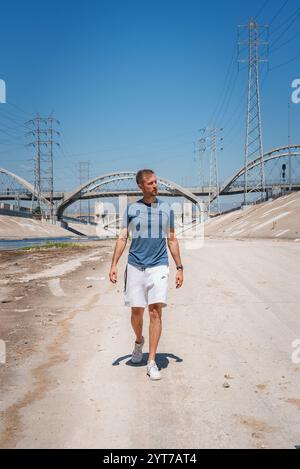 Un homme marche le long des rives bétonnées de la rivière Los Angeles. Le viaduc emblématique de la Sixth Street et des éléments industriels comme les lignes électriques sont visibles. Banque D'Images