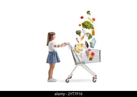 Petite fille avec un panier et des produits d'épicerie tombant dans le chariot isolé sur fond blanc Banque D'Images