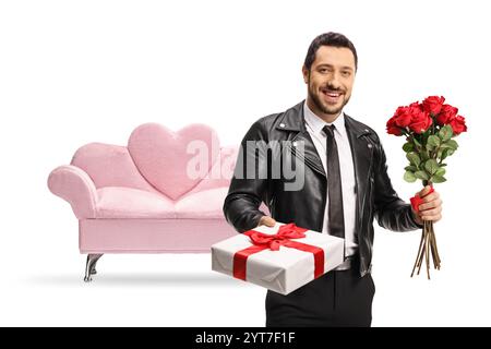 Homme dans une veste en cuir tenant une boîte cadeau et un bouquet de roses rouges devant un canapé en velours rose isolé sur fond blanc Banque D'Images