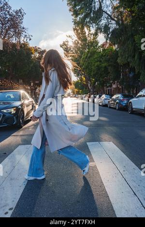 Une personne traverse un passage pour piétons dans une rue bordée d'arbres, avec des voitures garées et des guirlandes lumineuses. La lumière du soleil souligne le mouvement de leurs cheveux et de leur pelage. Banque D'Images