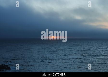 Le soleil se couche sur l'océan Pacifique près de Monterey, Californie, projetant une lueur orange à travers les nuages. De douces vagues ondulent sur l'eau calme. Banque D'Images