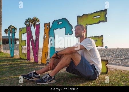 Un homme est assis sur l'herbe près d'un panneau vibrant Venice à Venice Beach, Los Angeles. Les palmiers, la plage de sable et le ciel bleu clair créent une Californie du Sud Banque D'Images