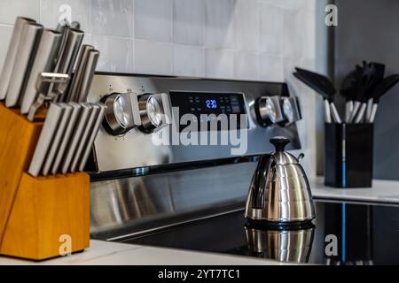 Cuisinière électrique en acier inoxydable avec dessus en verre, affichage numérique et une bouilloire posée sur la surface lisse de la table de cuisson. Un bloc de couteaux en bois et utensi Banque D'Images