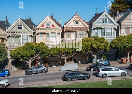 Les dames peintes, maisons victoriennes colorées, se dressent contre un ciel bleu clair à San Francisco. Les arbres et les voitures garées bordent le premier plan. Banque D'Images