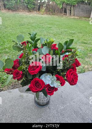 Un vase avec des roses rouges et un feuillage vert sur une table, placé sur un fond de pelouse verte et d'arbres, pris à la lumière du jour. Banque D'Images