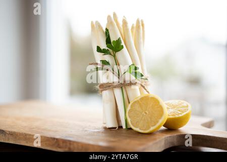 Bouquet debout d'asperges blanches fraîches. Légumes de printemps de saison avec persil et citron sur planche à découper en bois. Scène de cuisine pour la gastronomie saisonnière. Banque D'Images