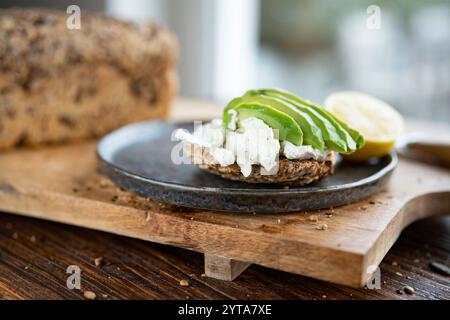 Tranches d'avocat fraîches sur pain aux grains avec fromage à la crème et citron. Nourriture végétarienne sur planche à découper en bois. Gros plan devant la fenêtre lumineuse. Faible profondeur de champ. Banque D'Images