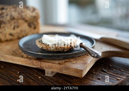 Pain frais à grains entiers avec fromage à la crème et assiette foncée sur une planche à découper en bois. Gros plan avec une faible profondeur de champ devant la fenêtre lumineuse. Banque D'Images