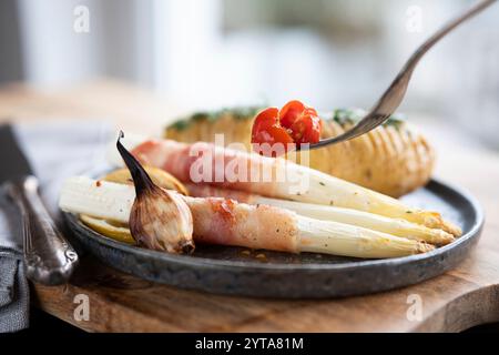 Asperges blanches frites enveloppées dans du bacon avec des pommes de terre aux herbes et des tomates sur une plaque de céramique noire courte profondeur de champ. Banque D'Images