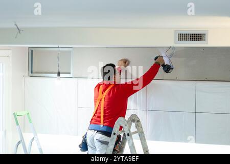 L'ouvrier en plaques de plâtre installe un mur en plaques de plâtre sur les armoires de cuisine pour couvrir le tuyau d'extraction de la hotte Banque D'Images