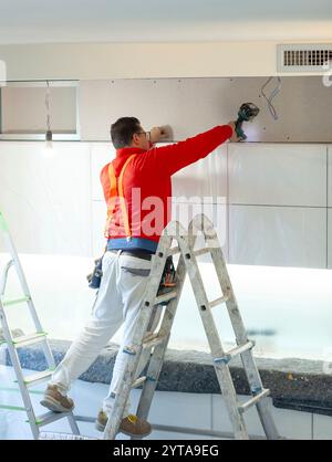 L'ouvrier en plaques de plâtre installe un mur en plaques de plâtre sur les armoires de cuisine pour couvrir le tuyau d'extraction de la hotte Banque D'Images