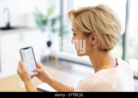 Femme caucasienne dans ses années 30 magasinant pour des cosmétiques sur le site de livraison en ligne à l'aide d'un smartphone. Elle est assise dans un environnement lumineux et moderne, se concentrant sur son écran de téléphone. Banque D'Images