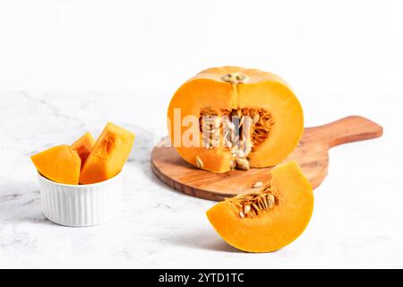 Une citrouille tranchée sur une planche à découper en bois rustique, reposant sur un comptoir de cuisine blanc propre, parfait pour les thèmes de cuisine d'automne. Banque D'Images