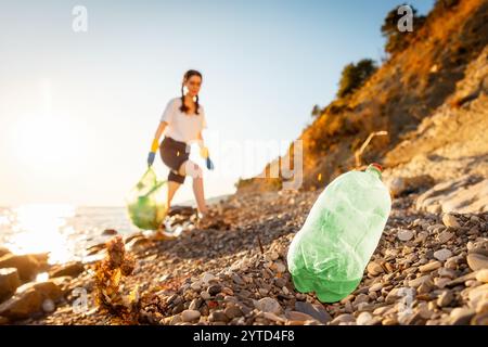 Vue défocalisée d'une femme bénévole dans des gants en caoutchouc tient un sac en plastique plein de déchets et marche vers le bas de la plage sauvage de galets. Gros plan de la bouteille en plastique i. Banque D'Images