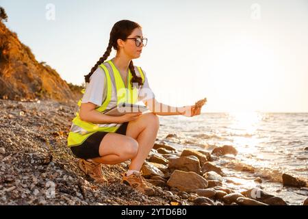 Jeune femme volontaire portant un gilet et tient une tablette électronique analysant le sol de la plage. En arrière-plan est l'océan, la côte et le coucher du soleil. Copier l'espace. Concept de Banque D'Images