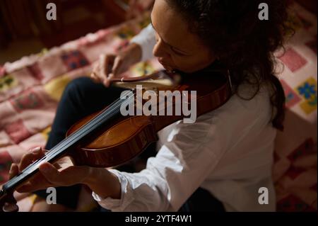 Une femme joue passionnément du violon assise sur une courtepointe colorée. Le cadre intime de la maison ajoute de la chaleur à l'instant musical. Banque D'Images