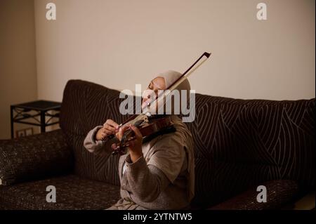 Une femme portant un hijab joue du violon, assise sur un canapé. Le cadre est calme et serein, mettant l'accent sur l'ambiance de détente et d'appréciation de la musique Banque D'Images