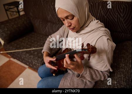 Une femme portant un hijab joue du violon assise sur un canapé confortable, se concentrant intensément sur sa pratique dans un environnement chaleureux et accueillant à la maison. Banque D'Images